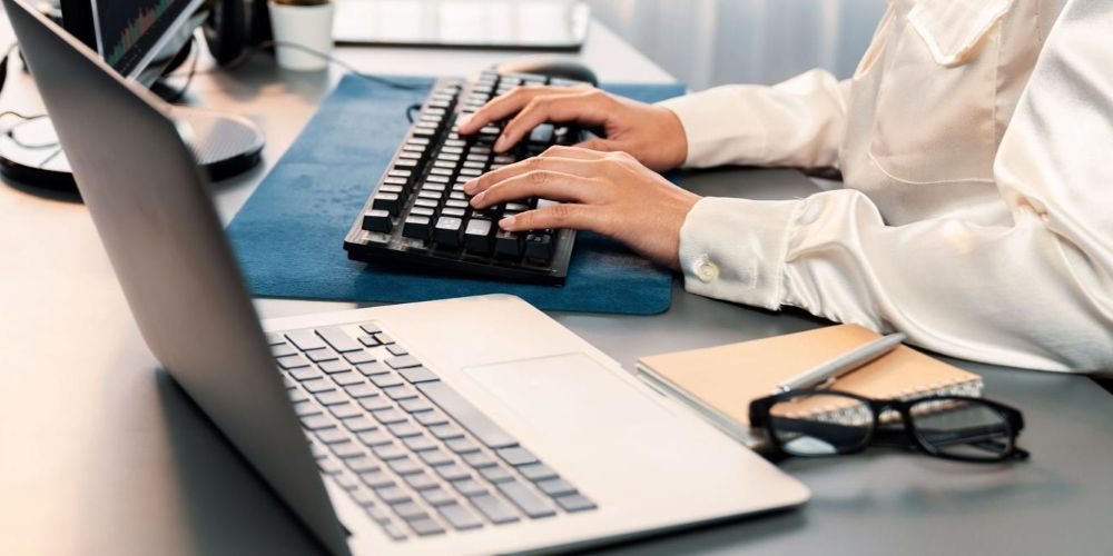 Na pierwszym planie otwarty laptop i okulary, w tle widać ręce osoby piszącej na klawiaturze komputerowej
