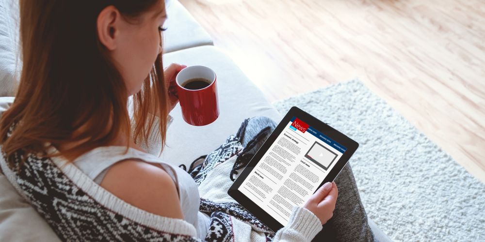 Kobieta przegląda treści w internecie na tablecie, w drugiej ręce trzyma kubek z kawą